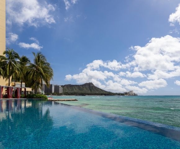 An infinity pool with blue water reflects palm trees and a resort building, overlooking a turquoise ocean with a distant mountain peak and scattered clouds.