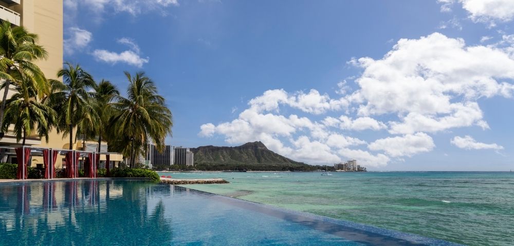 An infinity pool with blue water reflects palm trees and a resort building, overlooking a turquoise ocean with a distant mountain peak and scattered clouds.