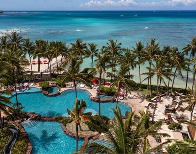 Aerial view of a tropical resort with multiple swimming pools, palm trees, lounge chairs, and a sandy beach with red umbrellas bordering a turquoise ocean.