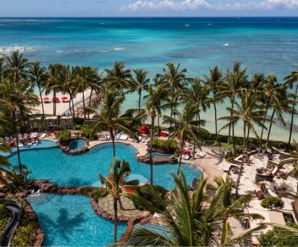 Aerial view of a tropical resort with multiple swimming pools, palm trees, lounge chairs, and a sandy beach with red umbrellas bordering a turquoise ocean.