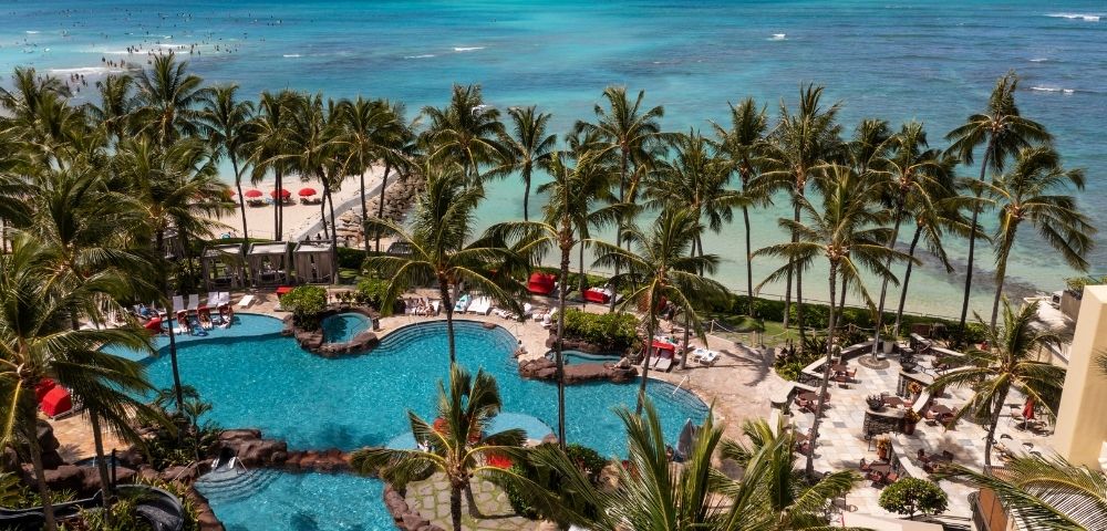 Aerial view of a tropical resort with multiple swimming pools, palm trees, lounge chairs, and a sandy beach with red umbrellas bordering a turquoise ocean.