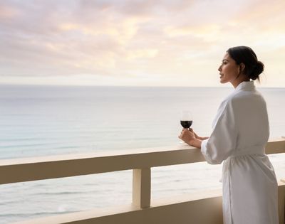 Woman in a white robe holding a glass of red wine on a balcony overlooking the ocean at sunset.