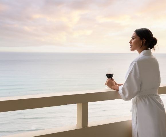 Woman in a white robe holding a glass of red wine on a balcony overlooking the ocean at sunset.