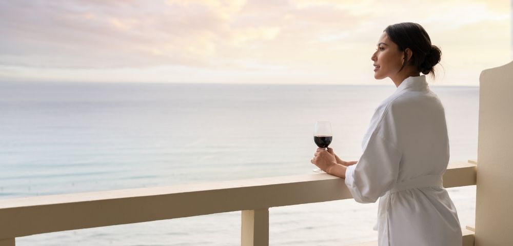 Woman in a white robe holding a glass of red wine on a balcony overlooking the ocean at sunset.