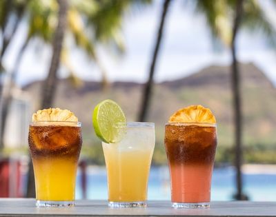 Three tropical cocktails with fruit garnishes are lined up on a table with a blurred background of palm trees, a mountain, and the ocean.