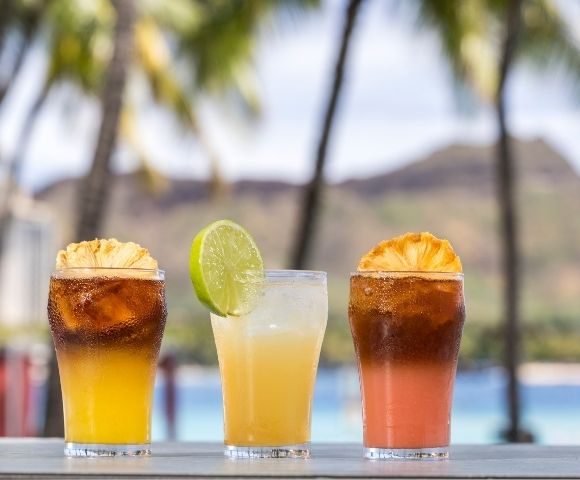 Three tropical cocktails with fruit garnishes are lined up on a table with a blurred background of palm trees, a mountain, and the ocean.
