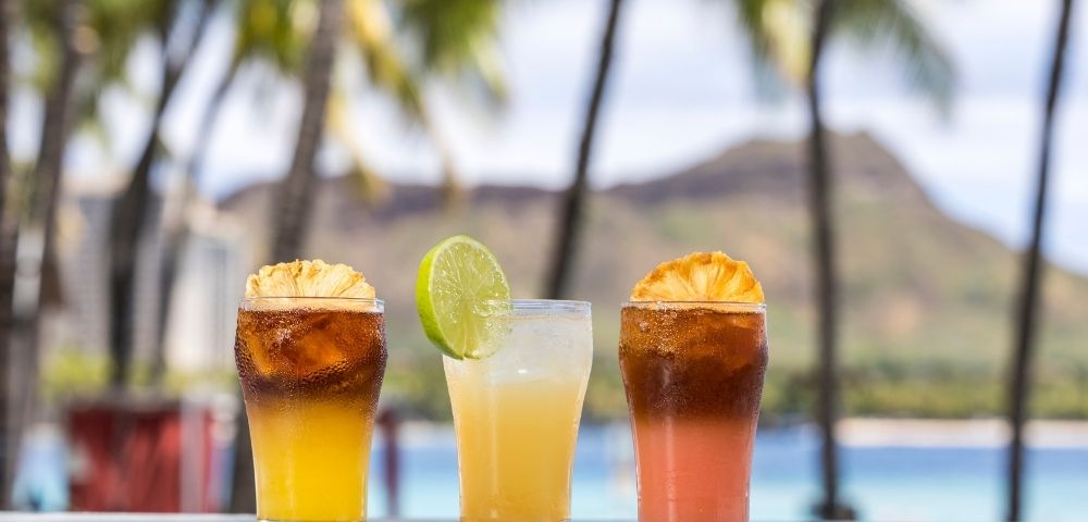 Three tropical cocktails with fruit garnishes are lined up on a table with a blurred background of palm trees, a mountain, and the ocean.