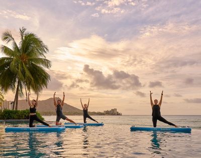 Four people practice yoga poses on inflatable paddleboards in a pool at sunset, with palm trees and ocean views in the background.