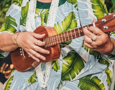 Close-up of hands playing a wooden ukulele, wearing rings and bracelets, against a backdrop of a blue and green tropical leaf-print shirt.