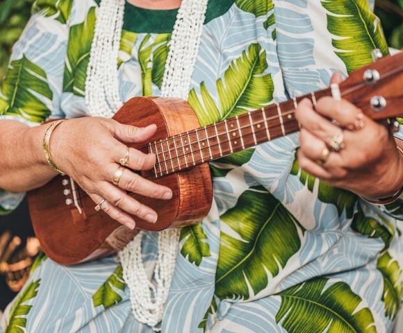 Close-up of hands playing a wooden ukulele, wearing rings and bracelets, against a backdrop of a blue and green tropical leaf-print shirt.