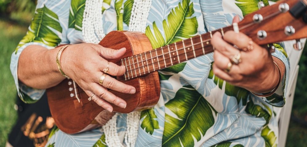 Close-up of hands playing a wooden ukulele, wearing rings and bracelets, against a backdrop of a blue and green tropical leaf-print shirt.
