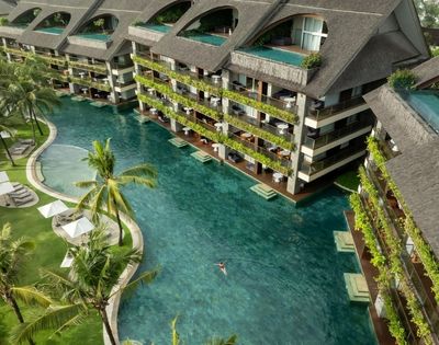 Aerial view of a resort with a large, clear turquoise pool winding between multi-story buildings with dark, pitched roofs and balconies overflowing with greenery; a lone swimmer is visible in the pool.