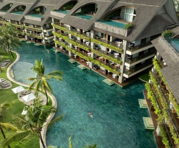 Aerial view of a resort with a large, clear turquoise pool winding between multi-story buildings with dark, pitched roofs and balconies overflowing with greenery; a lone swimmer is visible in the pool.