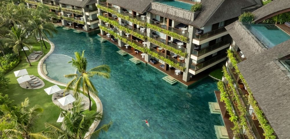 Aerial view of a resort with a large, clear turquoise pool winding between multi-story buildings with dark, pitched roofs and balconies overflowing with greenery; a lone swimmer is visible in the pool.