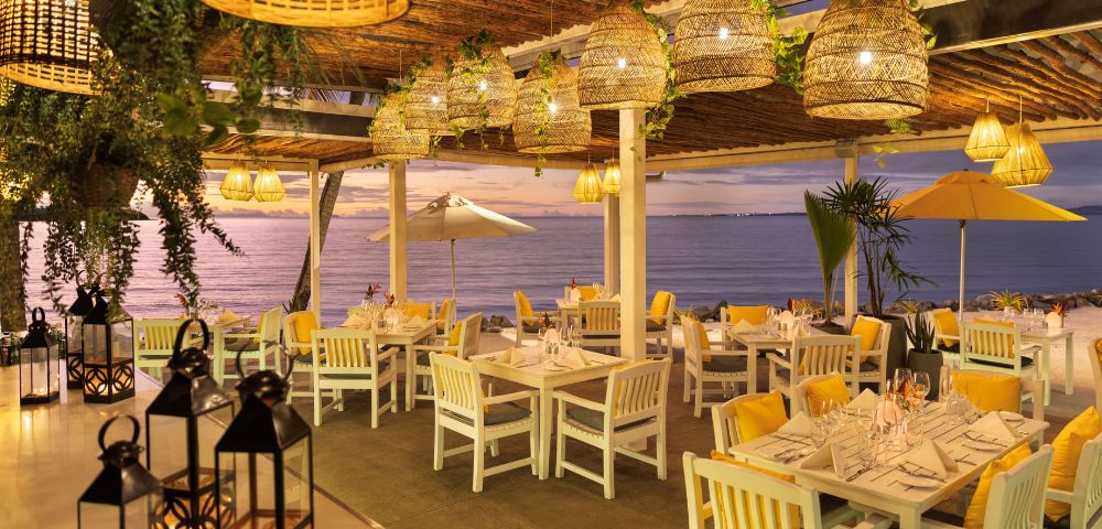 Seaside restaurant terrace at dusk, with white tables, woven pendant lights, lanterns, plants, and umbrellas overlooking a calm purple ocean.