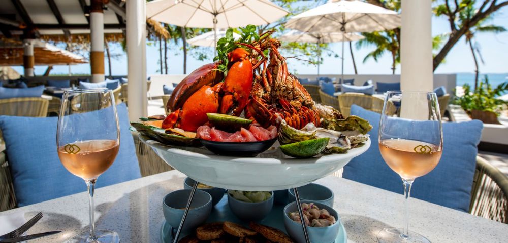 Lavish seafood platter with lobster and oysters sits between rosé wine glasses at a bright seaside restaurant, inviting and festive.