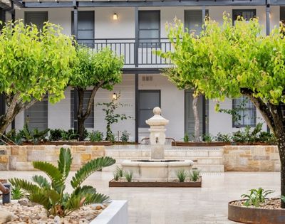 A classic stone fountain stands in a courtyard lined with trees and planters, in front of a white building with dark gray trim and a second-story balcony.