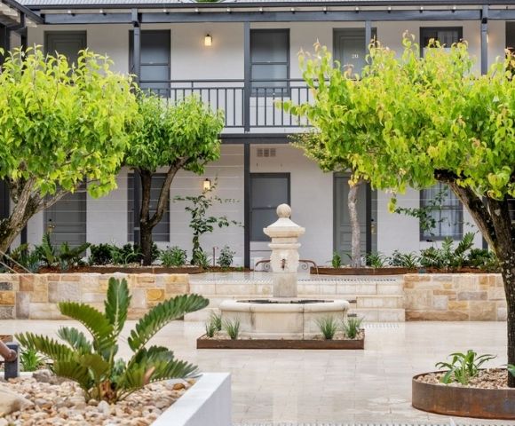 A classic stone fountain stands in a courtyard lined with trees and planters, in front of a white building with dark gray trim and a second-story balcony.