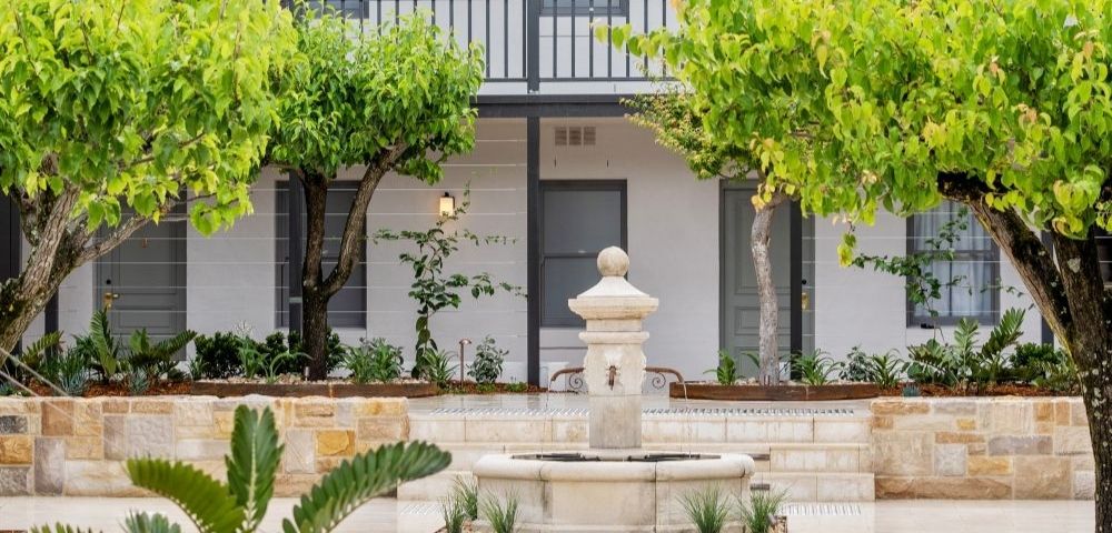 A classic stone fountain stands in a courtyard lined with trees and planters, in front of a white building with dark gray trim and a second-story balcony.