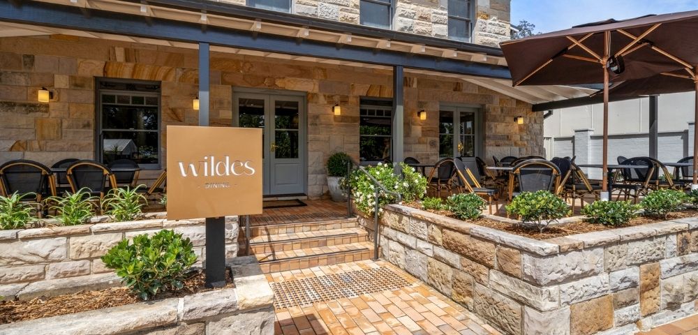 A stone building with a covered entrance and an outdoor dining area. A sign with 