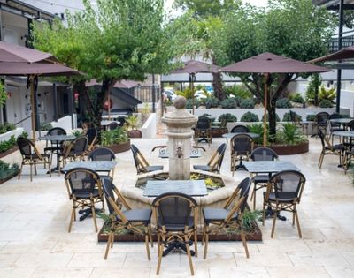 An outdoor courtyard cafe seating area with tables and chairs arranged around a stone fountain, under umbrellas and trees.