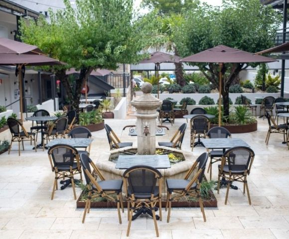 An outdoor courtyard cafe seating area with tables and chairs arranged around a stone fountain, under umbrellas and trees.