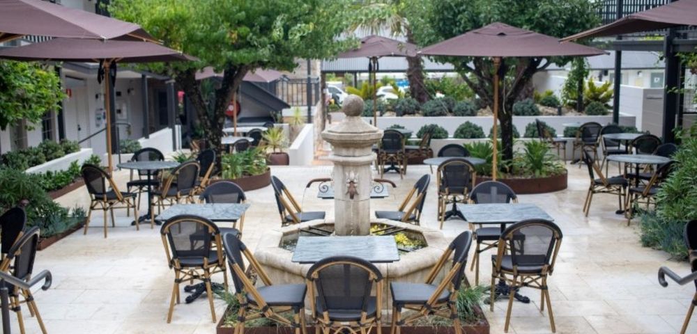 An outdoor courtyard cafe seating area with tables and chairs arranged around a stone fountain, under umbrellas and trees.