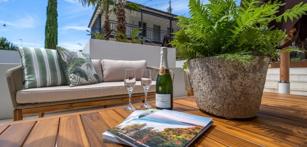 Outdoor patio setting with a sofa, coffee table, champagne bottle, two champagne flutes, a potted fern, and a brochure open on the table with a scenic landscape on its cover.
