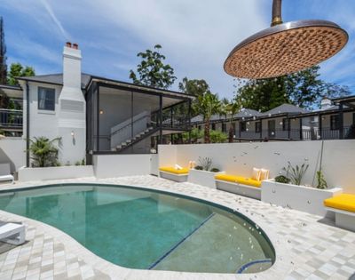 Outdoor swimming pool area with modern white building, yellow lounge chairs, and a large overhead showerhead with falling water droplets.