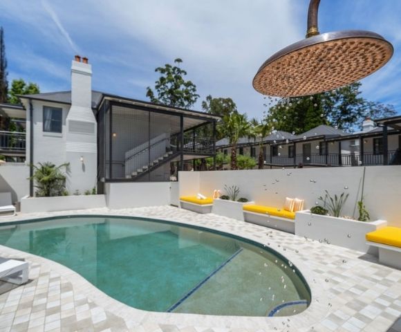 Outdoor swimming pool area with modern white building, yellow lounge chairs, and a large overhead showerhead with falling water droplets.