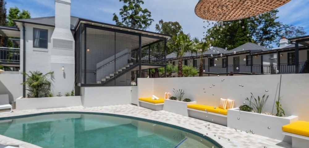 Outdoor swimming pool area with modern white building, yellow lounge chairs, and a large overhead showerhead with falling water droplets.