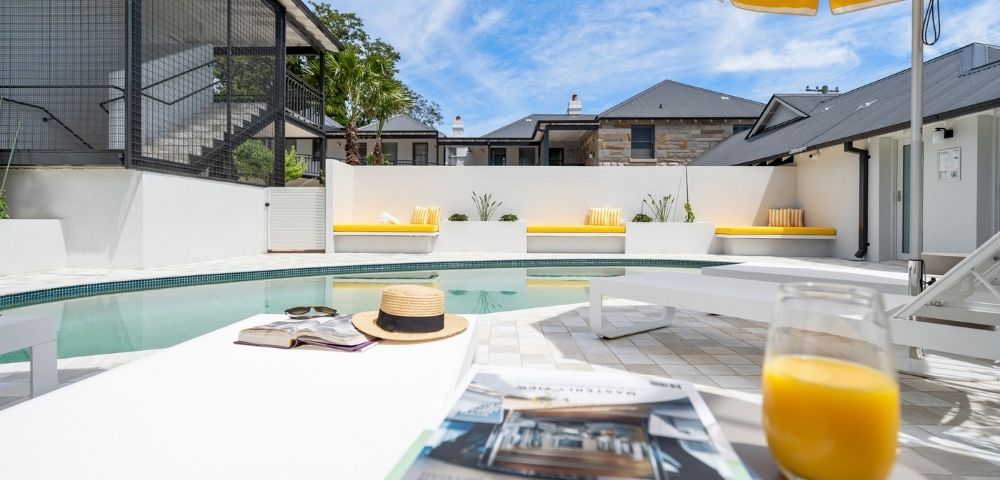 A bright, sunny day over a modern outdoor swimming pool area with yellow-cushioned lounge benches and a striped umbrella.
