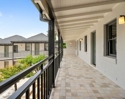 A long, covered outdoor walkway with a tiled floor and black railings, leading to doors of a building with white walls and dark-trimmed windows.