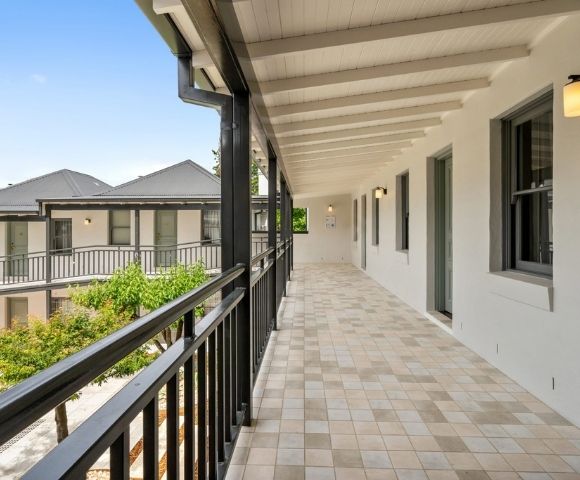 A long, covered outdoor walkway with a tiled floor and black railings, leading to doors of a building with white walls and dark-trimmed windows.