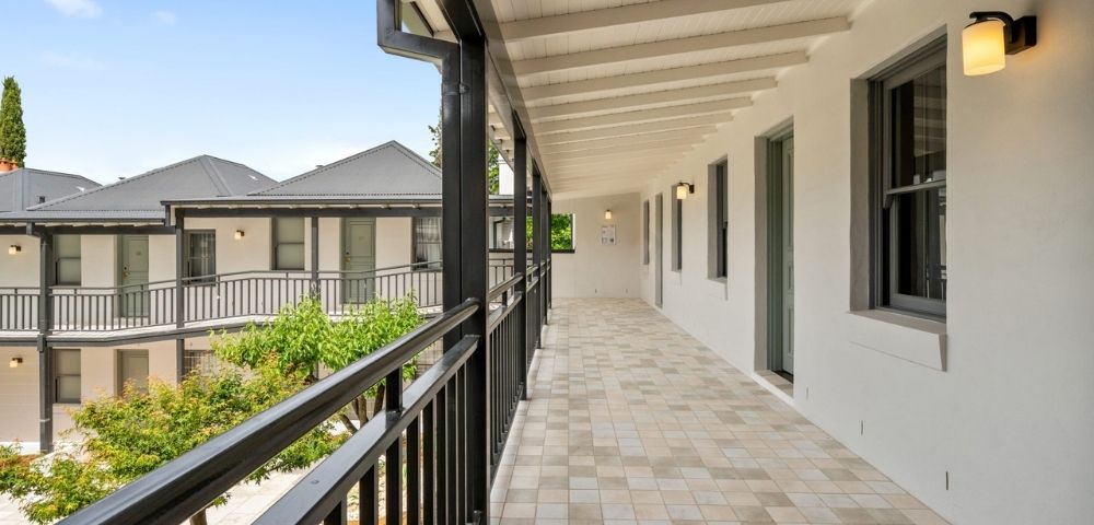 A long, covered outdoor walkway with a tiled floor and black railings, leading to doors of a building with white walls and dark-trimmed windows.