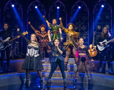 A diverse musical theater cast sings and dances on a blue-lit stage with guitars, drums, and dramatic costumes, creating an energetic mood.