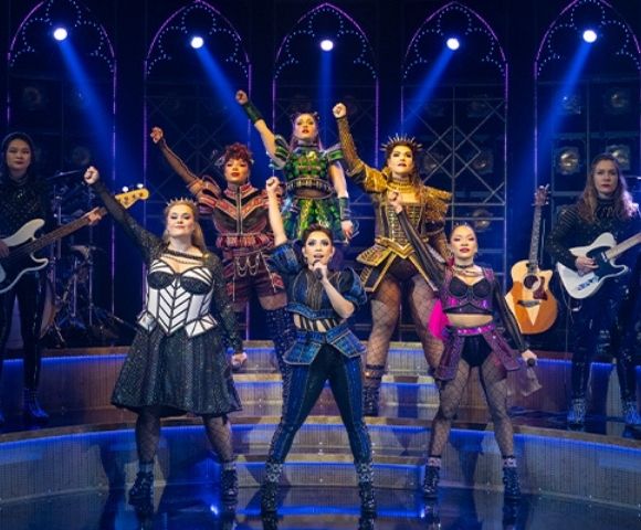 A diverse musical theater cast sings and dances on a blue-lit stage with guitars, drums, and dramatic costumes, creating an energetic mood.