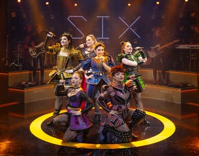 Six musical theater performers pose energetically onstage in colorful Tudor-inspired costumes, surrounded by musicians, amber lights, and a glowing yellow circle.