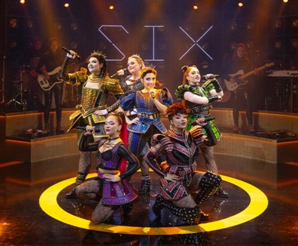 Six musical theater performers pose energetically onstage in colorful Tudor-inspired costumes, surrounded by musicians, amber lights, and a glowing yellow circle.