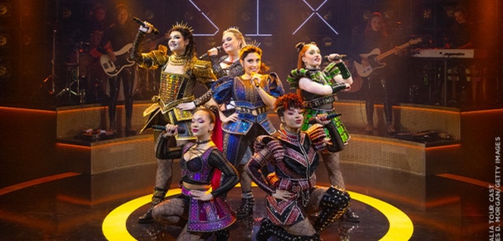 Six musical theater performers pose energetically onstage in colorful Tudor-inspired costumes, surrounded by musicians, amber lights, and a glowing yellow circle.