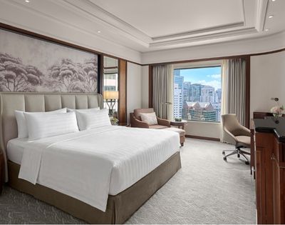 Elegant hotel bedroom with a neatly made king bed, Asian-inspired wall art, warm wood furnishings, and a bright city view through the window.