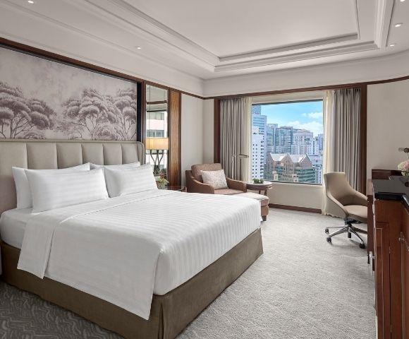 Elegant hotel bedroom with a neatly made king bed, Asian-inspired wall art, warm wood furnishings, and a bright city view through the window.