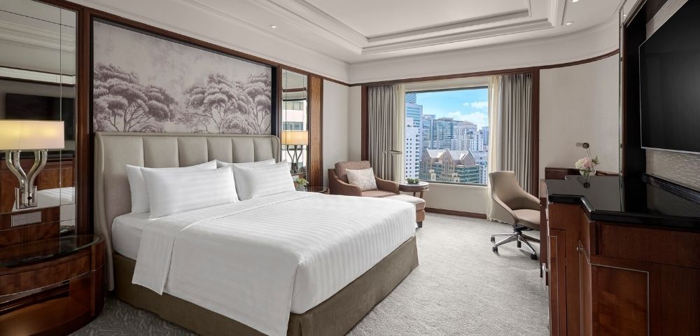 Elegant hotel bedroom with a neatly made king bed, Asian-inspired wall art, warm wood furnishings, and a bright city view through the window.