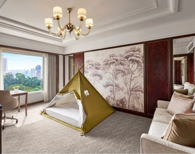 Elegant luxury hotel suite with a children’s teepee tent, cream sofa, chandelier, wall art, and sunny city view, blending comfort and playfulness.