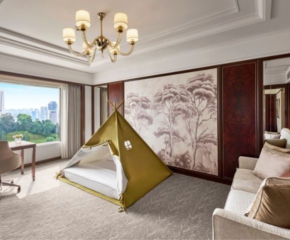 Elegant luxury hotel suite with a children’s teepee tent, cream sofa, chandelier, wall art, and sunny city view, blending comfort and playfulness.