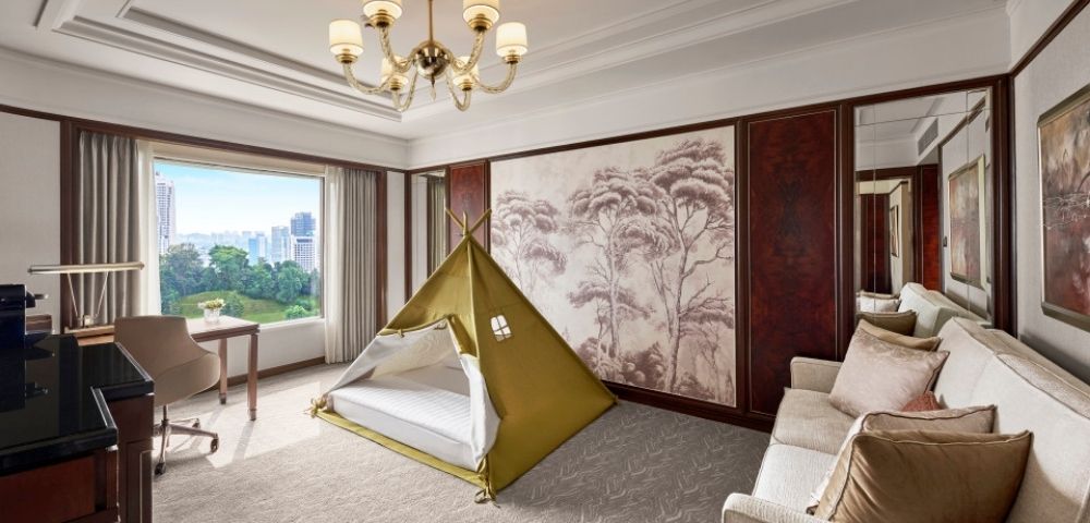 Elegant luxury hotel suite with a children’s teepee tent, cream sofa, chandelier, wall art, and sunny city view, blending comfort and playfulness.