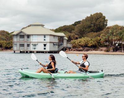 Two people paddle a green tandem kayak across calm water near a waterfront building and trees under cloudy skies, creating a relaxed scene.