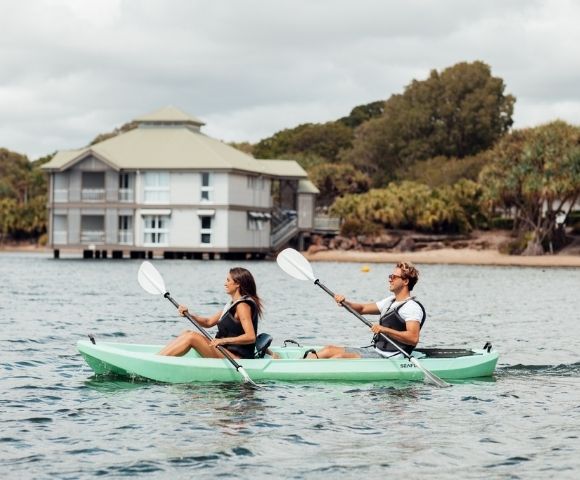 Two people paddle a green tandem kayak across calm water near a waterfront building and trees under cloudy skies, creating a relaxed scene.