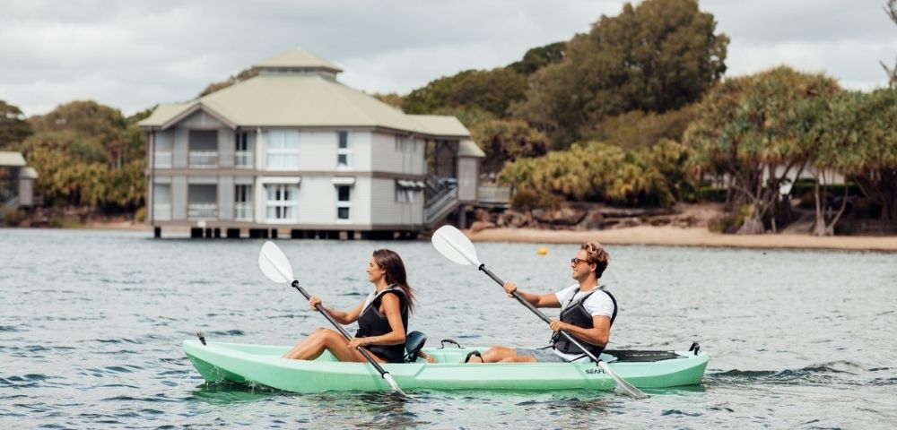 Two people paddle a green tandem kayak across calm water near a waterfront building and trees under cloudy skies, creating a relaxed scene.