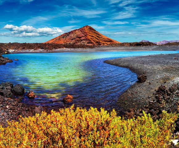 Panoramic view of a vibrant turquoise and green lagoon surrounded by dark volcanic rock and sparse yellow-orange vegetation, with a prominent red-brown mountain in the distance under a bright blue sky with scattered clouds.